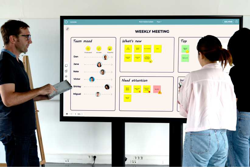Three people standing and discussing in front of a large digital screen displaying a weekly meeting board with team mood, updates, priorities, and attention points.