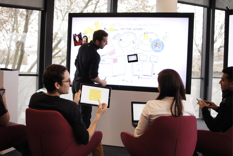Group of people in a meeting room brainstorming with a digital whiteboard and tablets, discussing creative ideas.
