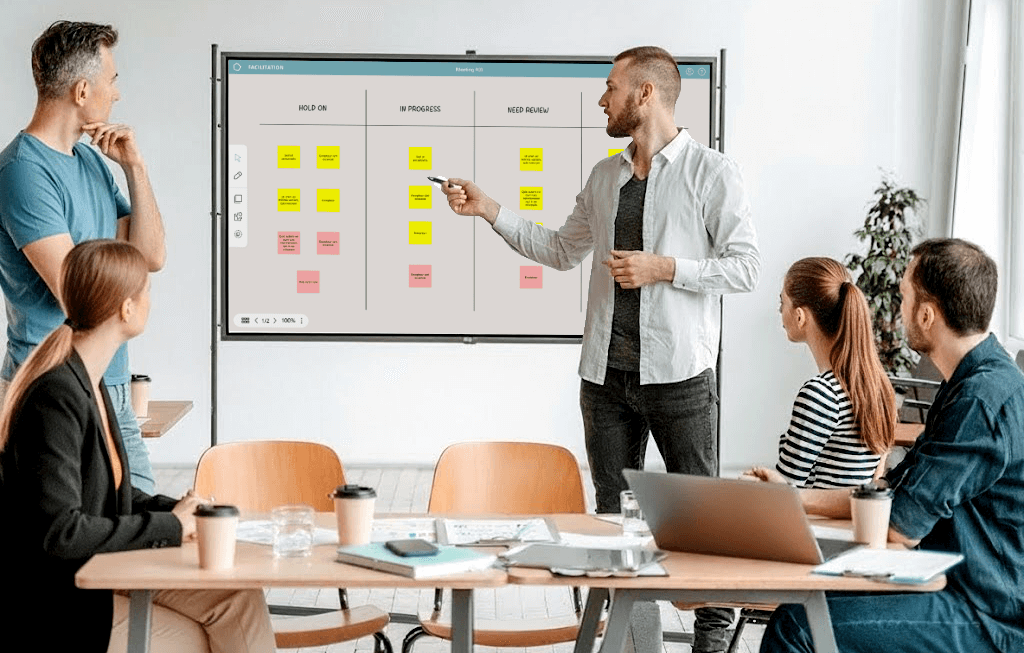 A man presenting to four colleagues in a meeting room, pointing at a digital board with columns labeled 'Hold On,' 'In Progress,' and 'Need Review' filled with color-coded sticky notes.