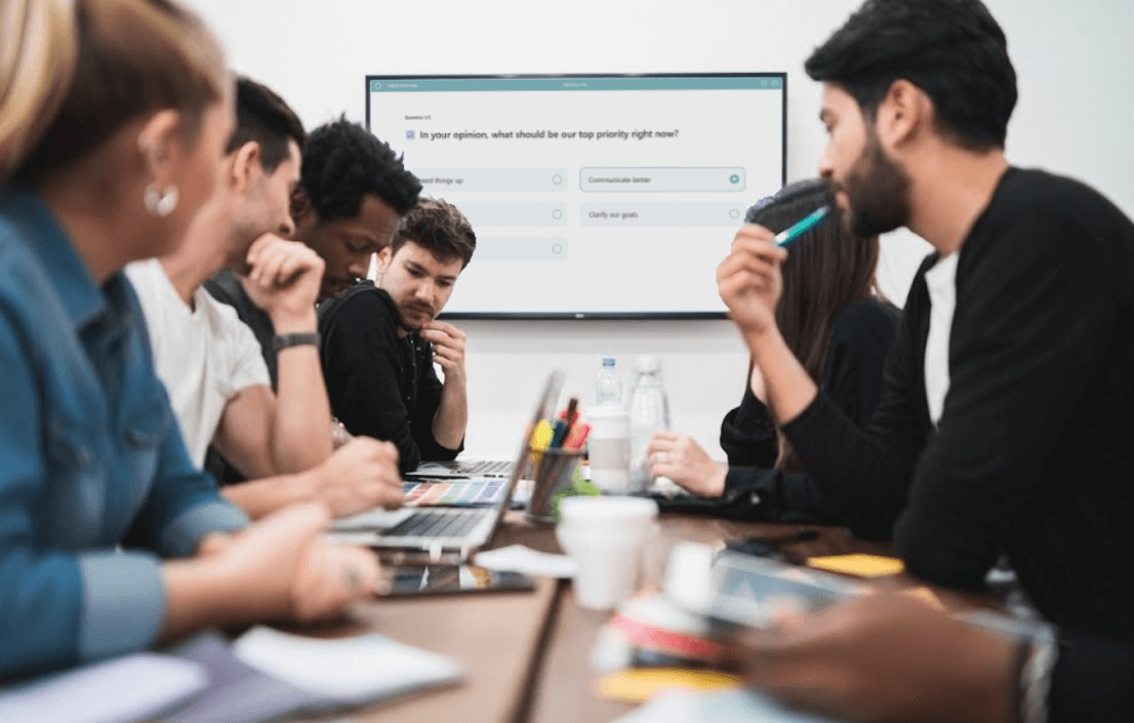 Group of people sitting around a table engaged in a meeting, looking at a screen displaying a question about top priority tasks.